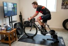Cyclist riding a smart trainer indoors with fan, mat, and training accessories set up around the bike